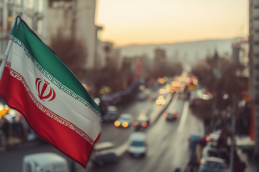 A vibrant Iranian flag stands out sharply against a soft-focus backdrop of city traffic and buildings, conveying a sense of national pride.